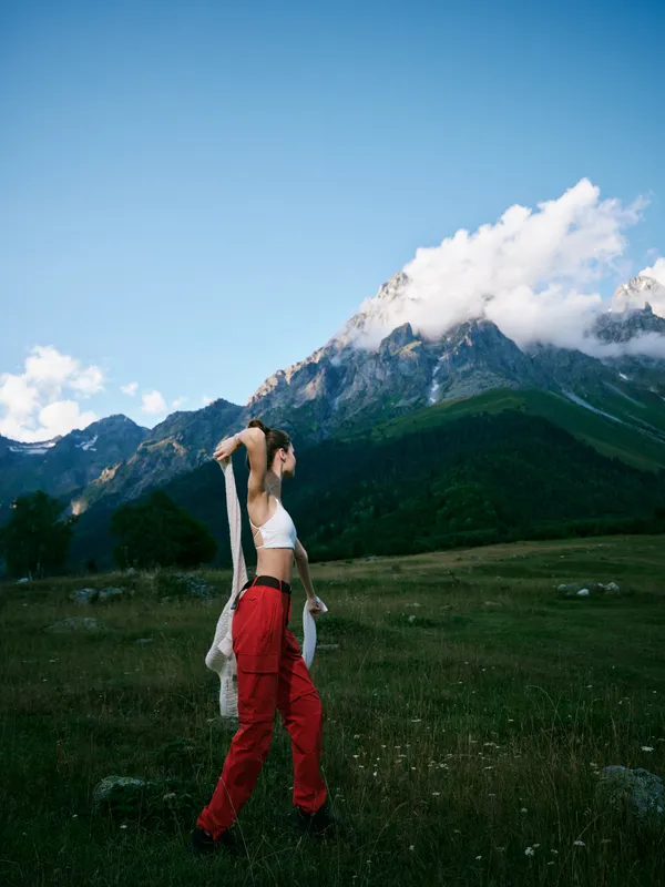 Hiker standing in red pants and white top in an alpine meadow, adjusting a jacket against the mountain backdrop, wide open landscape, clear sky, and sense of outdoor adventure