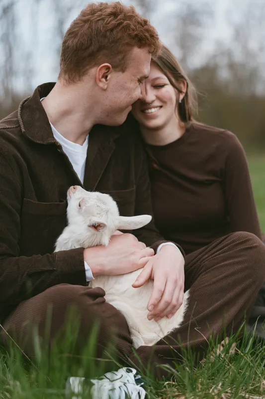 Young couple sitting on grass outdoors smiles warmly as the man gently holds a white baby goat in his arms during a relaxed lifestyle moment.