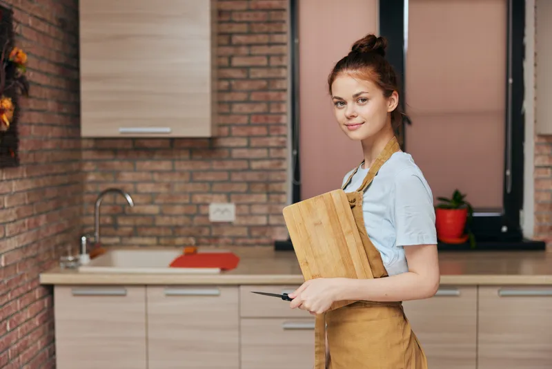 Весёлая женщина в коричневом фартуке режет разделочную доску на кухне