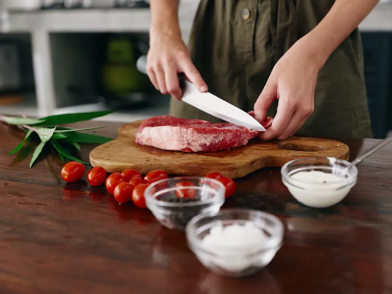 Женщина с ножом в руке нарезает свежее мясо для стейка для запекания на кухне