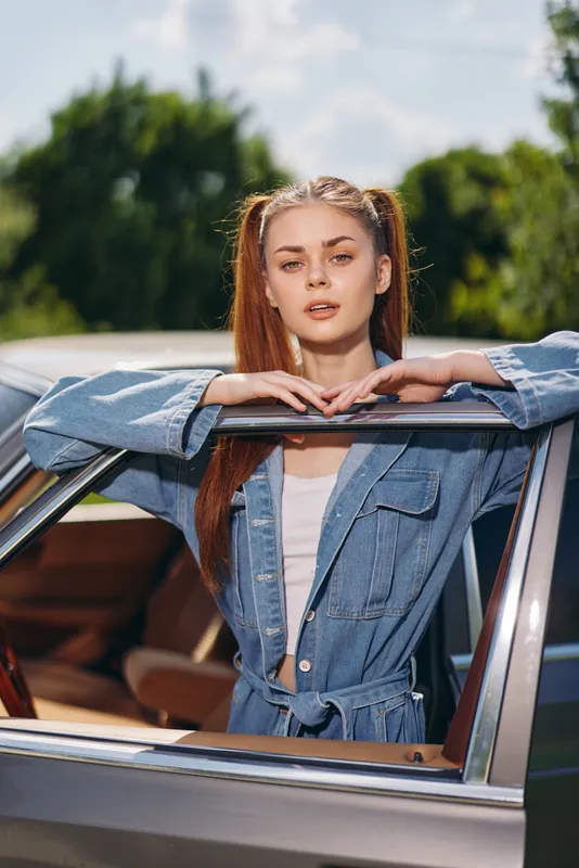 Young woman with long brown hair in denim jacket posing by vintage car, conveying confidence and youthfulness in a sunny outdoor setting.