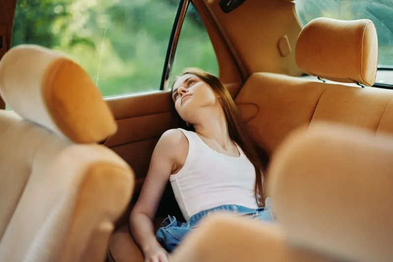 Young woman resting in a car, wearing a white tank top and denim shorts, exuding a peaceful and relaxed emotion against a warm natural background.