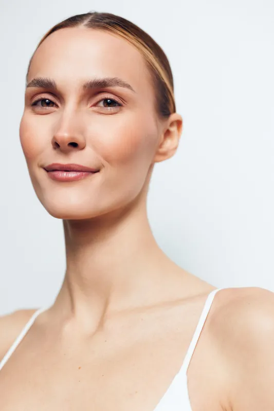 A confident young woman with clear skin and natural makeup looks directly at the camera. She wears a white tank top against a plain light background, symbolizing beauty and self-care.