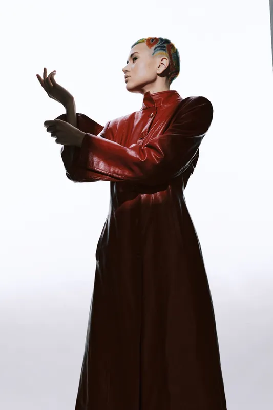 Woman with colorful hair wearing a long red leather coat, posing elegantly against a bright white background, showcasing modern fashion and unique style.