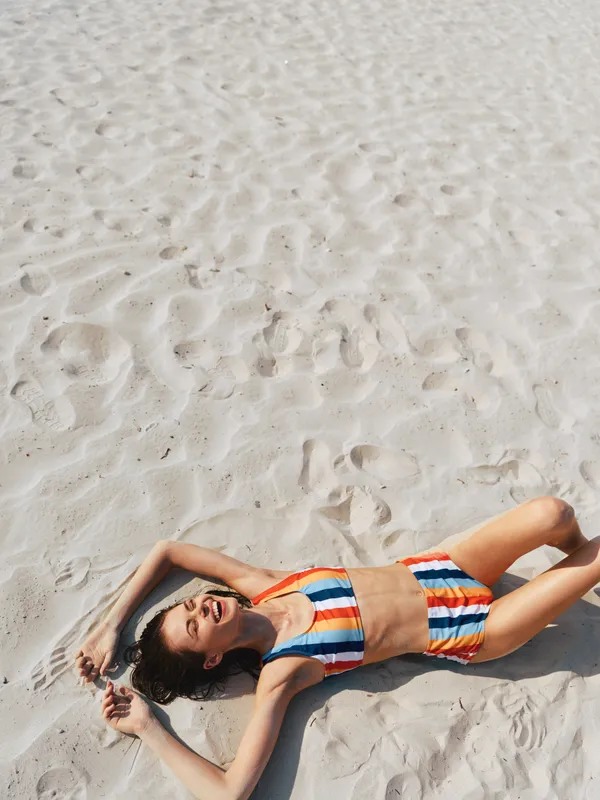 Young woman lying on white sand beach in a colorful striped bikini, sunlit and relaxed, capturing a carefree summer moment with warm tones and gentle breeze.