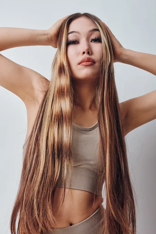 Young woman with long straight hair posing with hands behind head standing against neutral background. She wears casual sleeveless top and looks confidently at camera in studio.