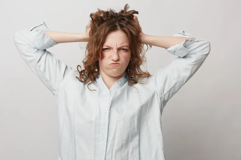 Frustrated woman with curly hair pulls her hair and frowns. She wears a white shirt and expresses stress and anger on a plain light background. Concept of frustration and tension.