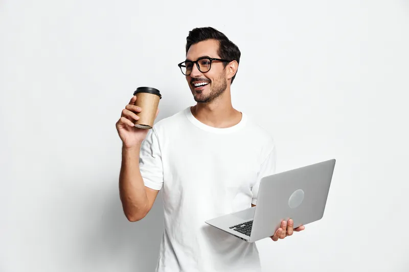 Cheerful smiling young man wearing glasses holding coffee cup and laptop while standing against plain backdrop. Modern lifestyle and work concept.