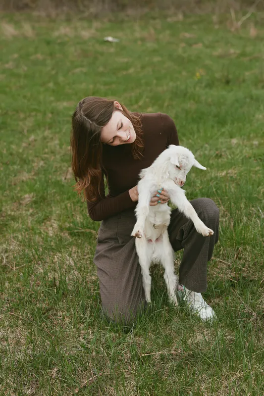 Young woman in brown sweater kneels on grass holding a white baby goat while smiling gently in a natural outdoor lifestyle scene.