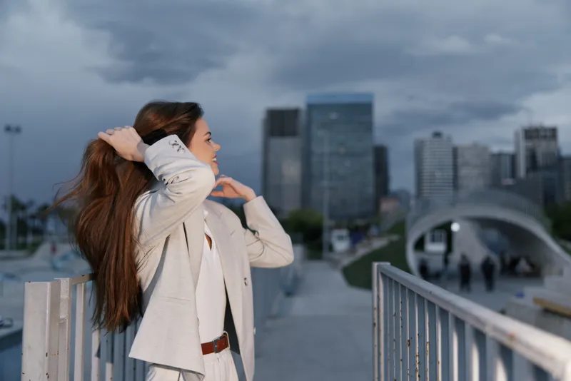 business woman in beige suit outdoors adjusting hair with city skyscrapers background under cloudy sky urban fashion style professional confident mood
