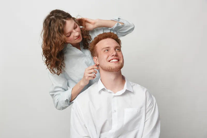 Smiling redhead man in white shirt sits while woman with curly hair adjusts his earring. Studio portrait on light background. Concept of friendship, care, and casual fashion accessories.