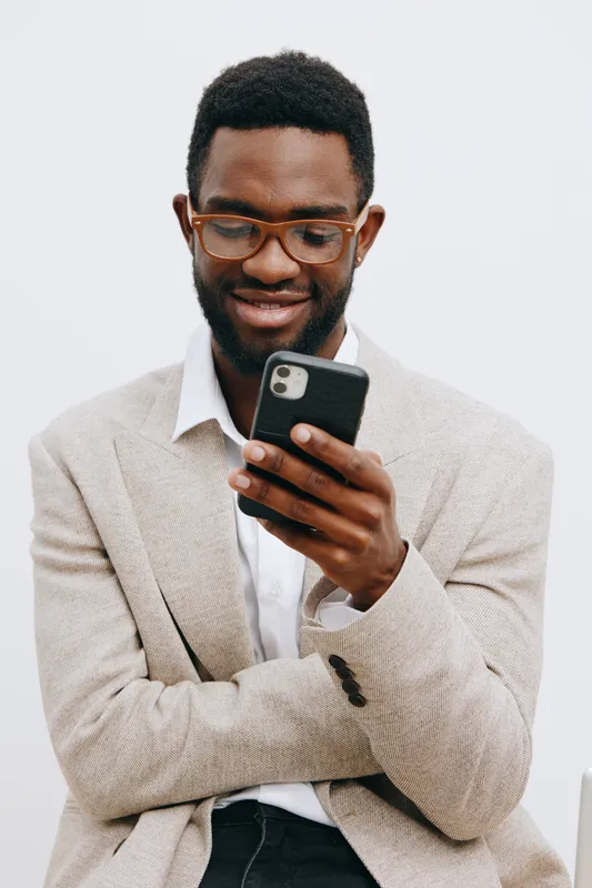 Smiling man using smartphone, casual beige blazer, glasses, dark skin, short curly hair, modern interior background, lifestyle concept, technology, communication, happiness, adult male, portrait, shot