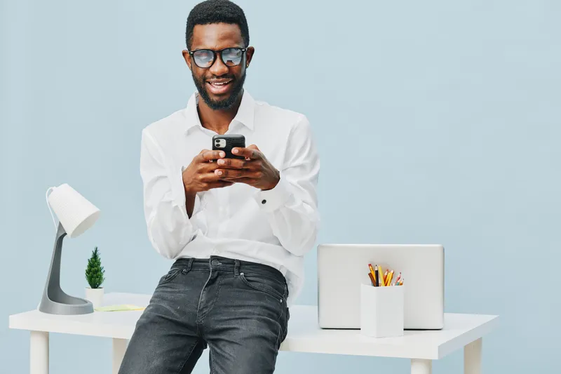 Smiling man using mobile phone in modern office setting, dressed casually in white shirt and dark jeans, with laptop, desk lamp, and stationery on plain light blue background. People lifestyle concept