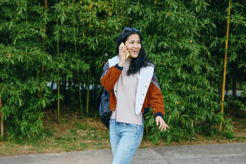 woman, phone, outdoors, casual, jacket, lifestyle, backpack, greenery, smile, street, communication, mobility, urban, nature, candid, photography, fashion, accessory, daylight, travel