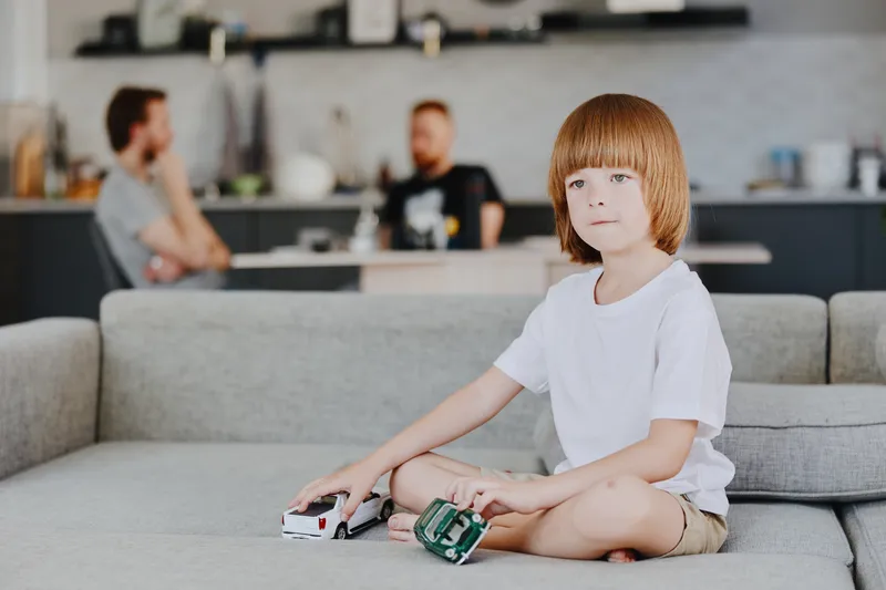 A thoughtful boy playing with toy cars in a cozy living room, surrounded by adults. The serene atmosphere captures a moment of childhood joy and imagination.