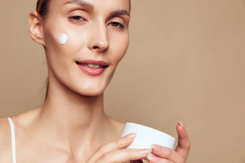 Young woman with short hair applies moisturizing cream on her cheek while holding a white jar against a beige background with a calm expression.