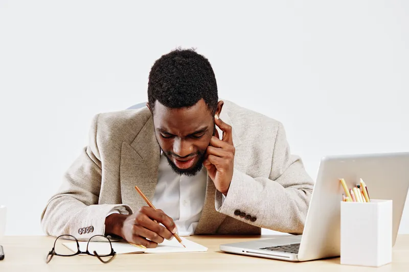 Professional young African American man in beige blazer working at desk with laptop and notepad, having phone conversation, smiling, in modern office setting, professional lifestyle concept.