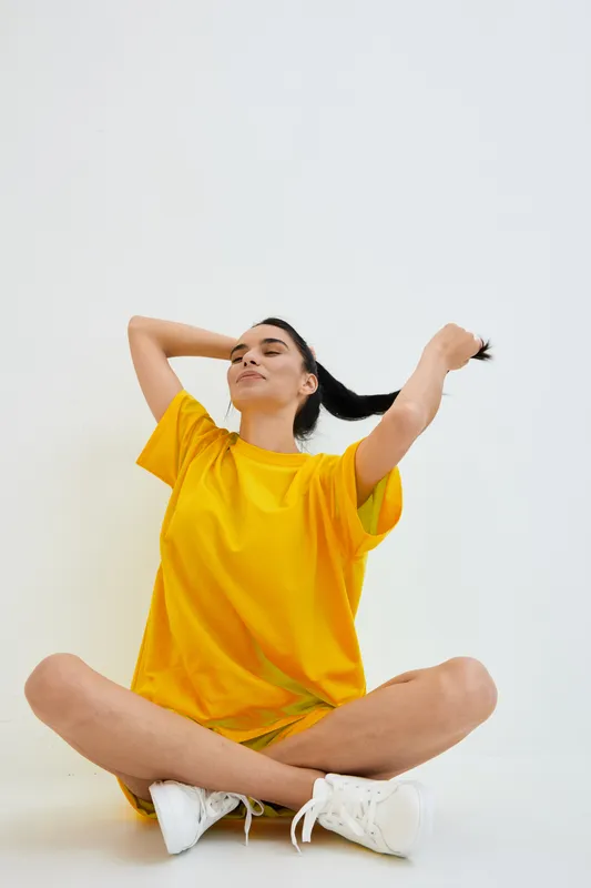 young woman wearing bright yellow shirt sitting cross-legged on white background, casual style, relaxed pose, playful mood, indoor lighting, minimalistic setting