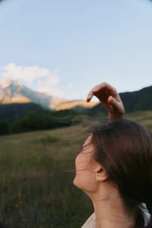Young woman outdoors raises a gentle hand in a light breeze, standing in a grassy field with distant mountains under a clear sky, a calm sunlit mood