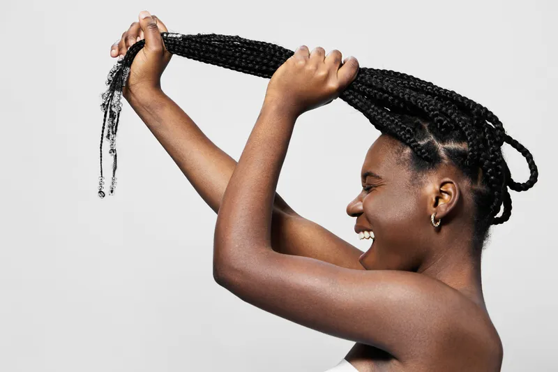 Joyful woman with braided hair showcasing her hairstyle, emphasizing beauty and confidence in a minimalist setting with a light background