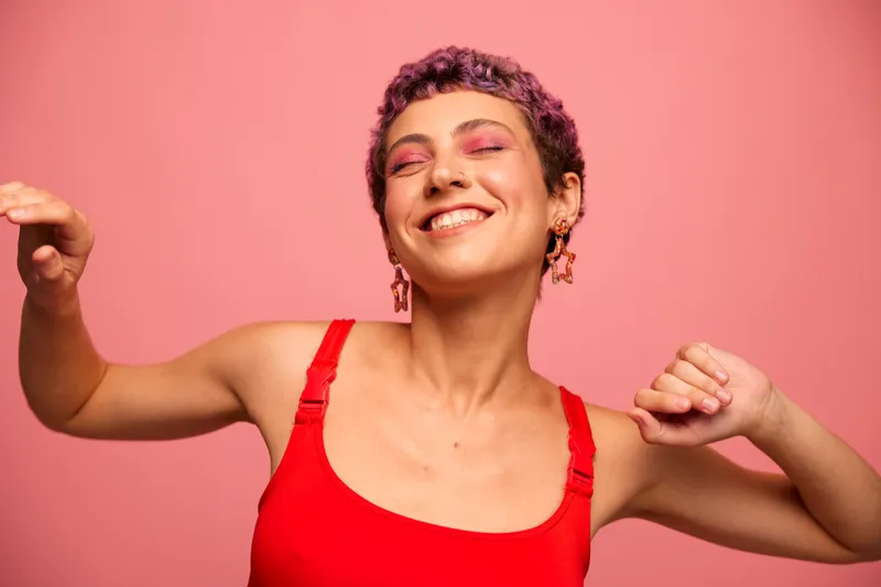 Модный портрет женщины с короткой стрижкой фиолетового цвета и улыбкой с зубами в красном топе на розовом фоне, которая счастливо танцует