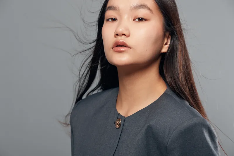 Portrait of confident Asian young woman with long hair wearing gray formal jacket standing against gray background. Expression serious and professional, fashion and beauty concept.