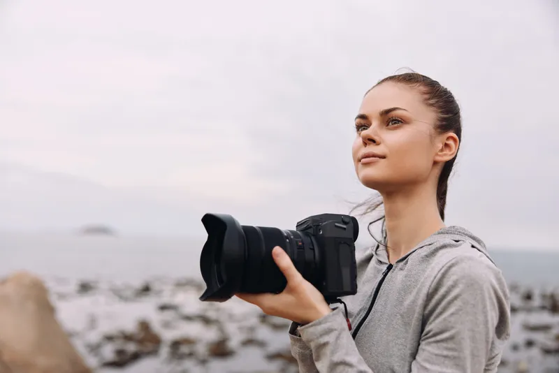 A young woman with an Eastern European appearance holding a DSLR camera, smiling and enjoying the coastal scenery, reflecting passion for photography and nature