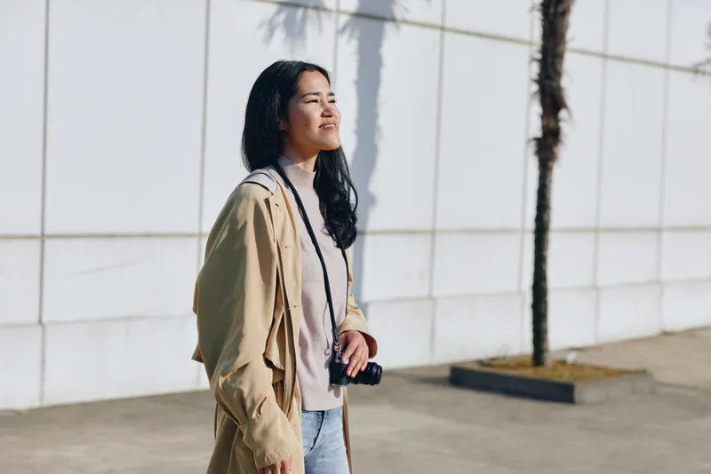 Smiling woman with a camera in hand walks outdoors beside a white wall, casual fashion and sunlight glow create a candid moment of urban exploration and daily adventure