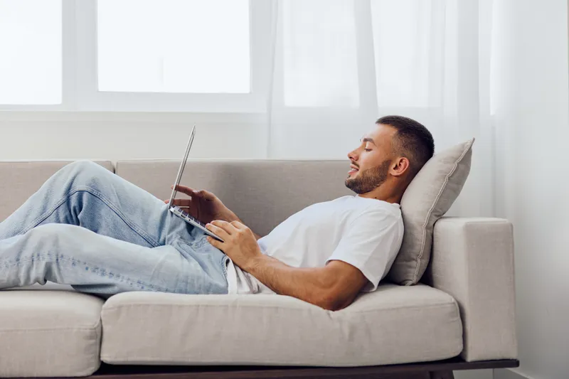 Relaxed man using a laptop, enjoying comfortable indoor lifestyle. Bright, modern space evoking tranquility and productivity. Perfect for remote work themes.