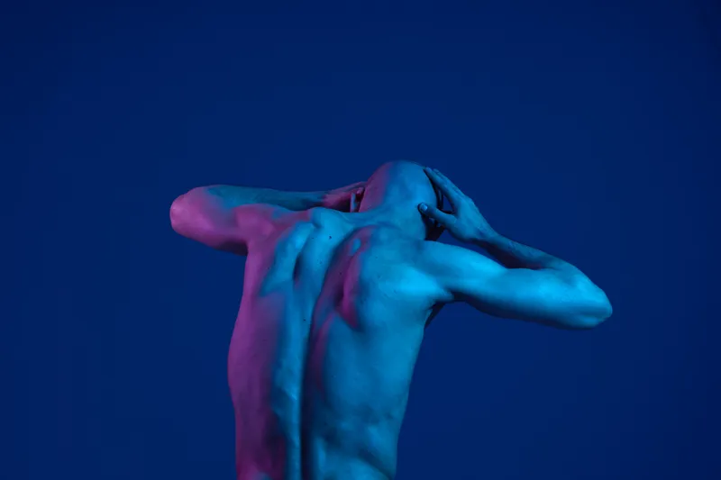 A man poses dramatically against a blue background, showcasing his muscular physique with vibrant lighting. The image emphasizes emotion and artistic expression, creating a striking visual impact.