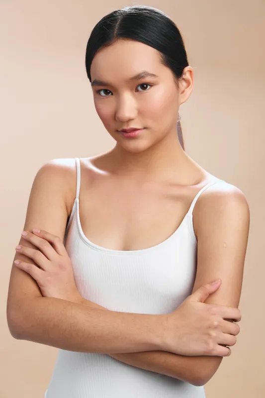 asian woman stands in a neutral studio setting with arms crossed, wearing a white tank top. she gazes softly at the camera, conveying confidence and calm in a clean portrait.