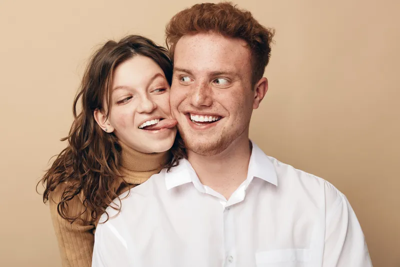 Playful young woman with curly hair sticks out tongue behind smiling redhead man in white shirt. Casual couple having fun together on beige background. Concept of friendship and joy.