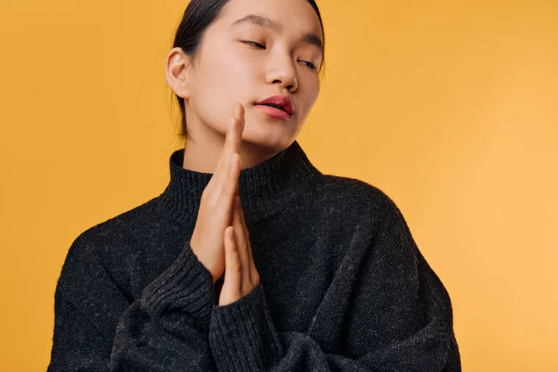 Woman in black sweater with eyes closed and hands together on orange background for meditation, relaxation, mindfulness, calmness, serenity, wellness, peace and self care.