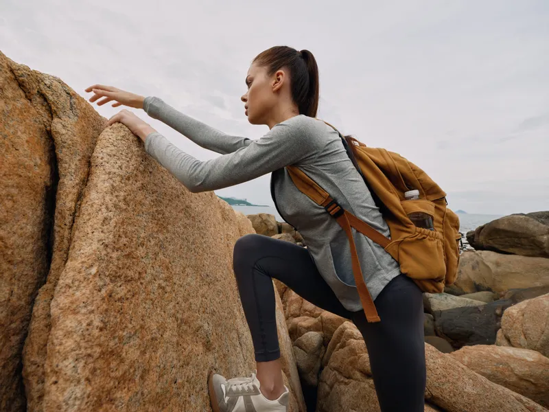 Young female climber with a backpack climbing rocks against a cloudy sky, showcasing adventure, determination, and a spirit of exploration