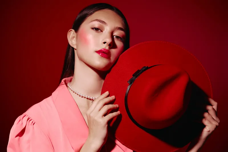 Fashionable young woman with glowing skin wears glossy red makeup and round pearl necklace, holding vibrant red wide-brim hat against a vivid red background in a beauty creative concept.
