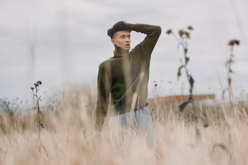 A young man stands alone in tall dry grass wearing a knit sweater, photographed in candid natural light to convey genuine authenticity and calm contemplative presence with subtle muted tones.