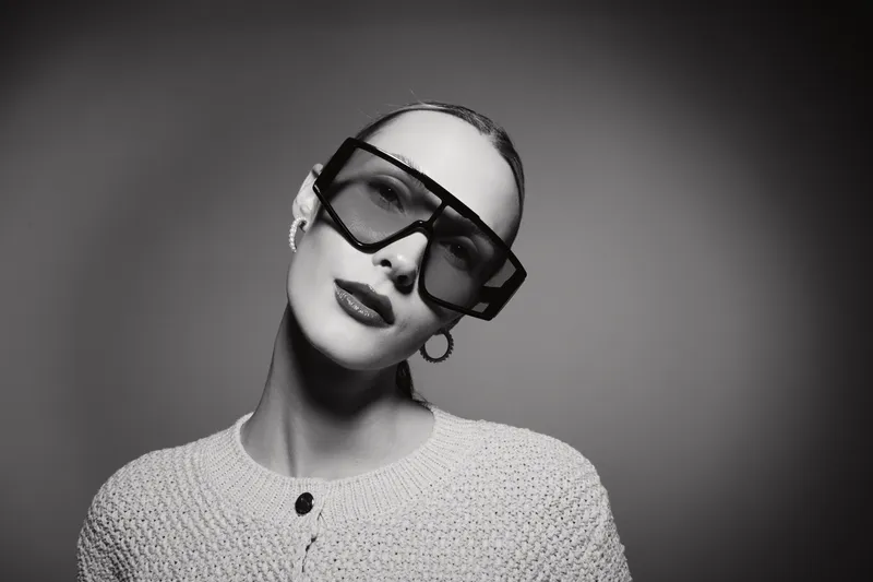 Elegant white woman with pale skin and slicked back hair wears large black sunglasses and knitted sweater posing in studio. Concept of fashion, style, and modern portrait.