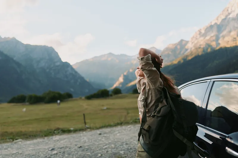Woman lifestyle traveler standing near car in mountain valley, stretching arms, enjoying fresh air, casual outfit, sunny day, scenic nature background