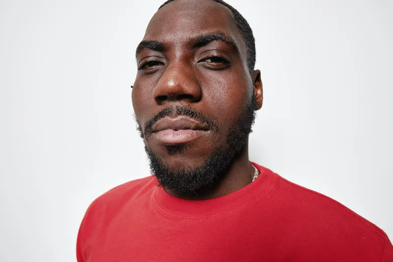 Portrait of confident black man with dark skin wearing red shirt looking at camera with slight smile on white background. Closeup, facial hair and casual style with natural light.