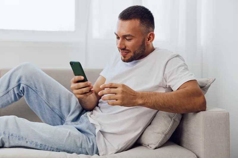 Relaxed man using smartphone while sitting on a cozy couch, wearing casual clothing, with a cheerful expression, capturing the essence of modern lifestyle and technology.