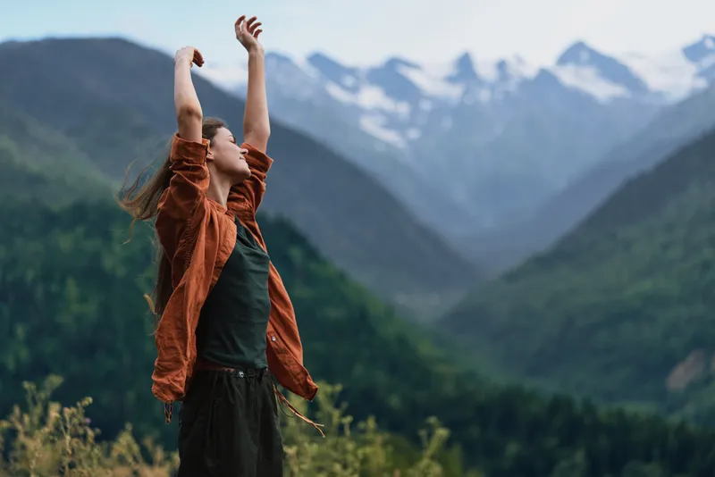 Joyful woman raises arms in a mountain landscape, wearing an orange jacket, expressing freedom and adventure in a wide outdoor setting showing freedom