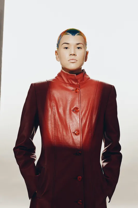 Young woman with colorful short hair wearing a stylish red leather jacket, standing confidently against a plain light background, modern fashion portrait.
