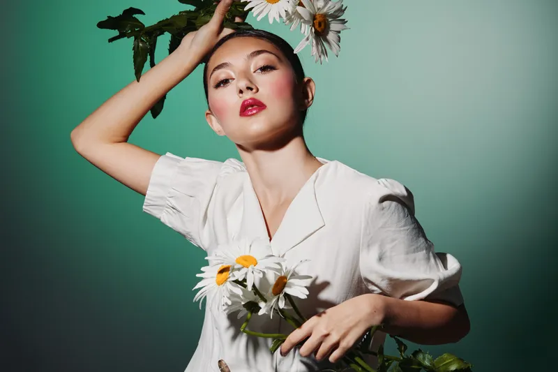 Fashionable young woman wearing white blouse holds daisies against green background. Her bright makeup and pose create a fresh beauty and creative fashion concept for editorial shoots.
