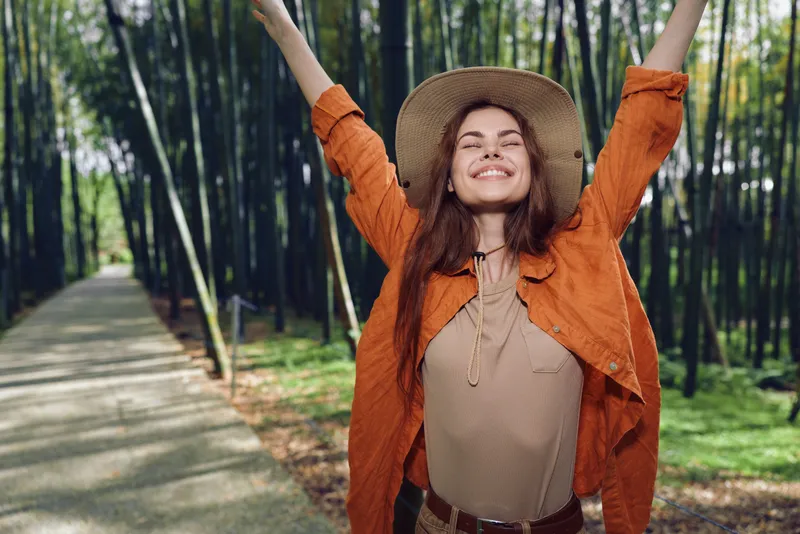 Woman with smile and straw hat standing on forest path, arms raised in joyful nature moment. Young traveler in orange jacket enjoys sunlight, freedom and outdoor happiness and bamboo grove