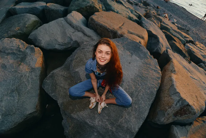 Lifestyle woman in modern boho-western denim street style sitting on rocks near water, smiling with natural red hair and film color tone at sunset outdoors.