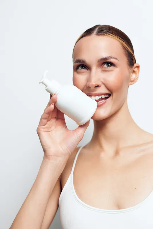 Young woman with brunette hair uses a white pump bottle for skincare, smiling in a bright clean studio with a minimal, fresh moment.