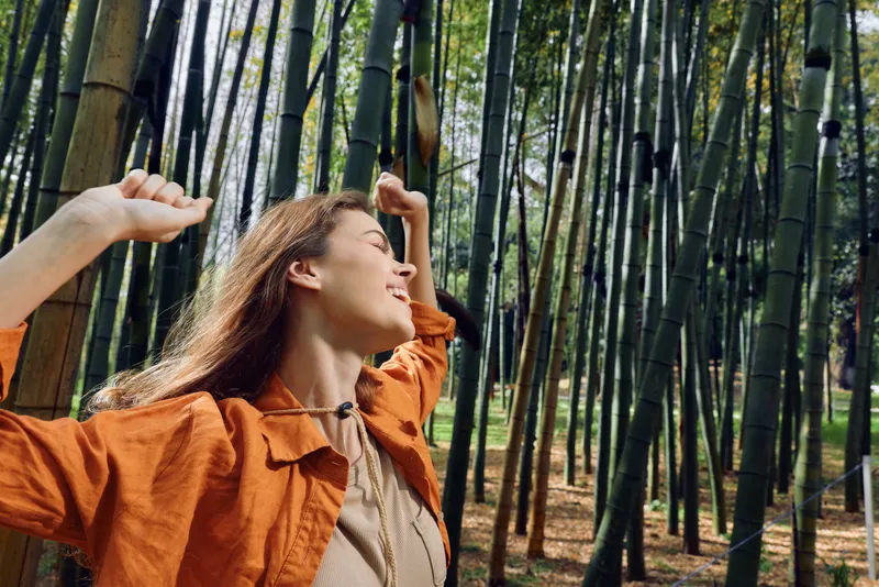 Woman in orange jacket bamboo forest portrait showing nature relaxation and joy, eyes closed and arms raised; outdoor lifestyle with sunlight filtering through tall green bamboo trees.