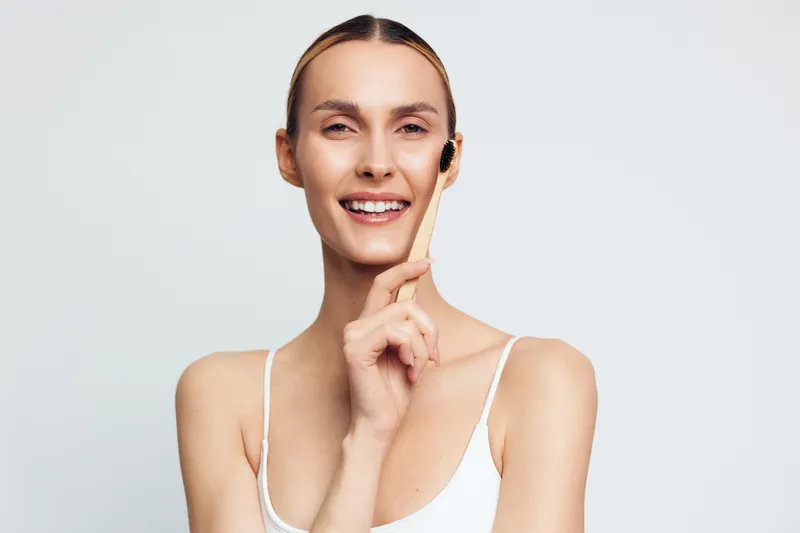 Smiling young woman with clear skin holds facial brush near cheek in skincare routine isolated on white background, demonstrating beauty and personal care concept with natural expression.