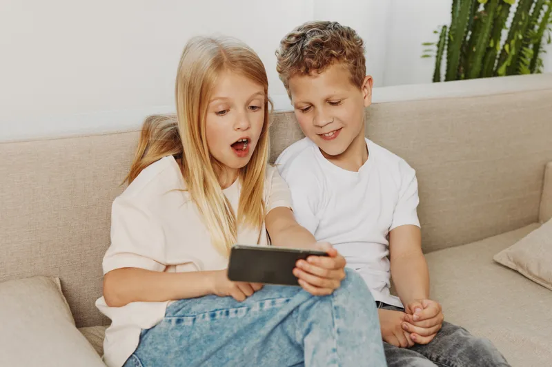 Two curious children sitting on sofa watching smartphone with surprised and happy expressions in bright cozy living room environment.