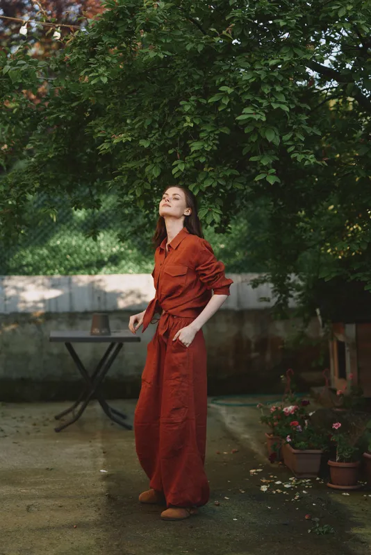 Cheerful woman outdoors in a rust red dress, standing with relaxed posture under shaded trees, sunlight filtering through leaves, natural colors, garden background, serene lifestyle moment, candid